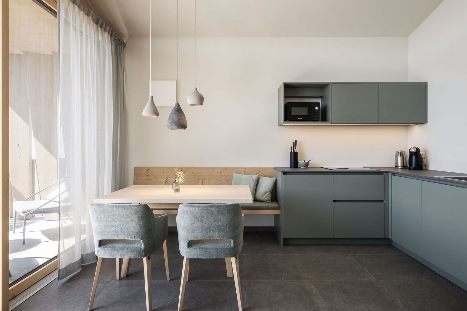 Moderne, minimalistische Küche mit grünen Schränken, Esstisch und zwei Stühlen vor einem Fenster mit weißen Vorhängen.