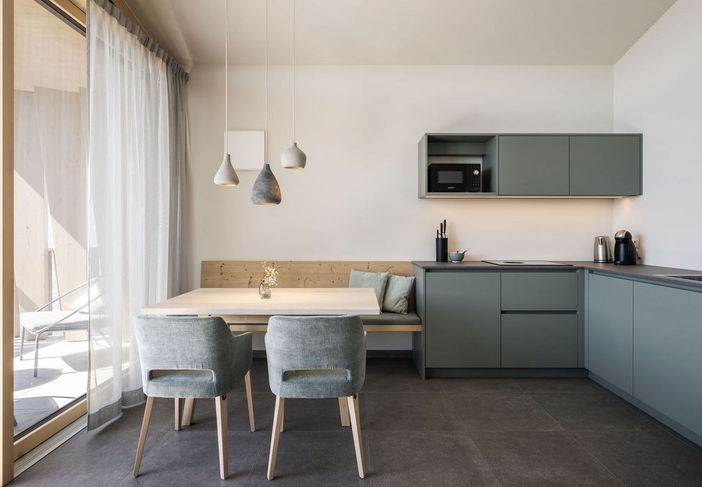 Moderne, minimalistische Küche mit grünen Schränken, Esstisch und zwei Stühlen vor einem Fenster mit weißen Vorhängen.