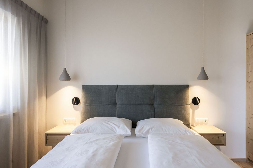 Modernes Schlafzimmer mit grauem Kopfteil, zwei weißen Kissen und Bettdecken, zwei Nachttischen mit Hängelampen und Vorhängen am Fenster.