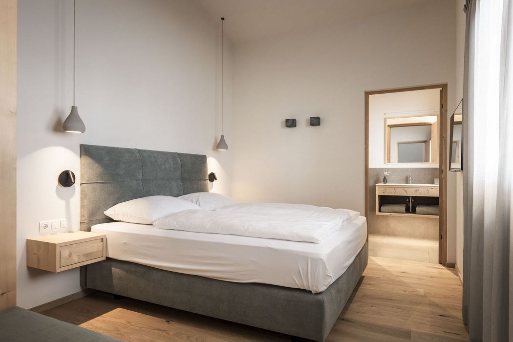 Modernes, helles Schlafzimmer mit Doppelbett, grauem Polsterbett, hängenden Lampen und offenem Türrahmen zum Badezimmer