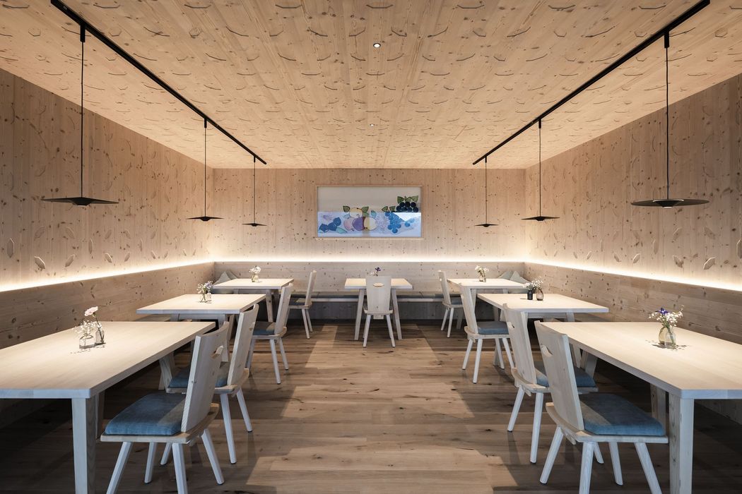 Moderner, minimalistisch eingerichteter Raum mit Holzvertäfelung, Holztischen und -stühlen, dezenter Beleuchtung und Blumenvasen auf den Tischen.