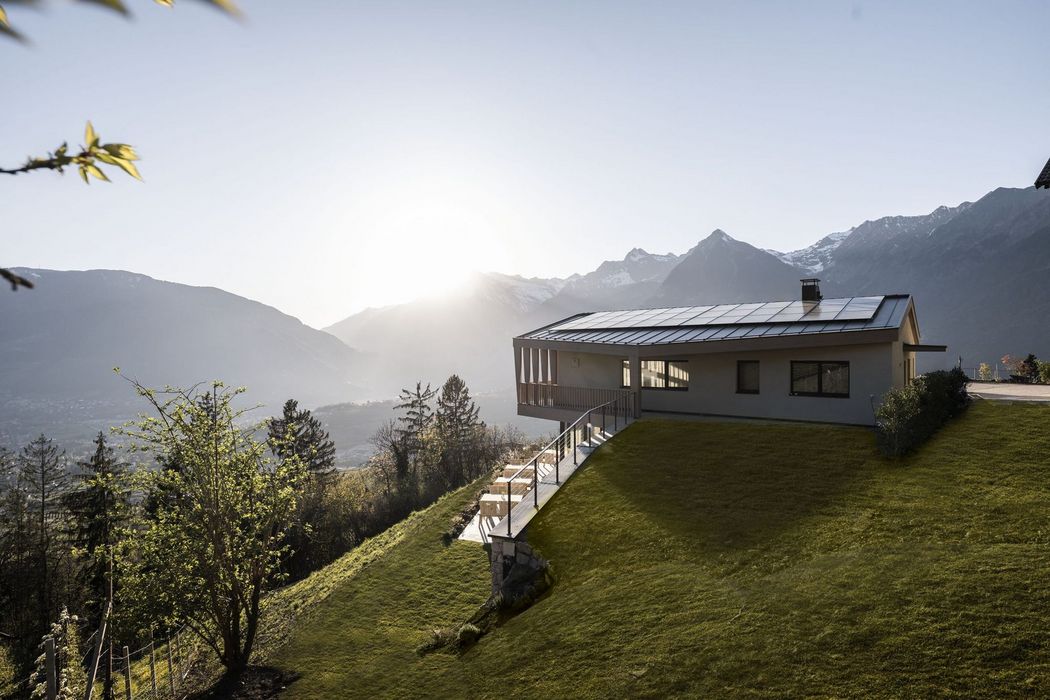 Modernes Haus mit Solarpanelen auf dem Dach in hügeliger Landschaft mit Sonnenuntergang und Bergen im Hintergrund