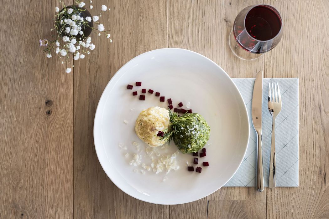 Zwei knödelartige Speisen, eine grün und eine hell, garniert mit kleinen roten Würfeln und Kräutern auf einem weißen Teller, neben Messer, Gabel und Glas Rotwein auf Holztisch.