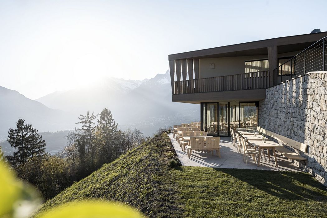 Moderner Außenbereich eines Hauses mit Holzmöbeln auf einer Terrasse, umgeben von Hügeln und Bergen im Sonnenlicht.