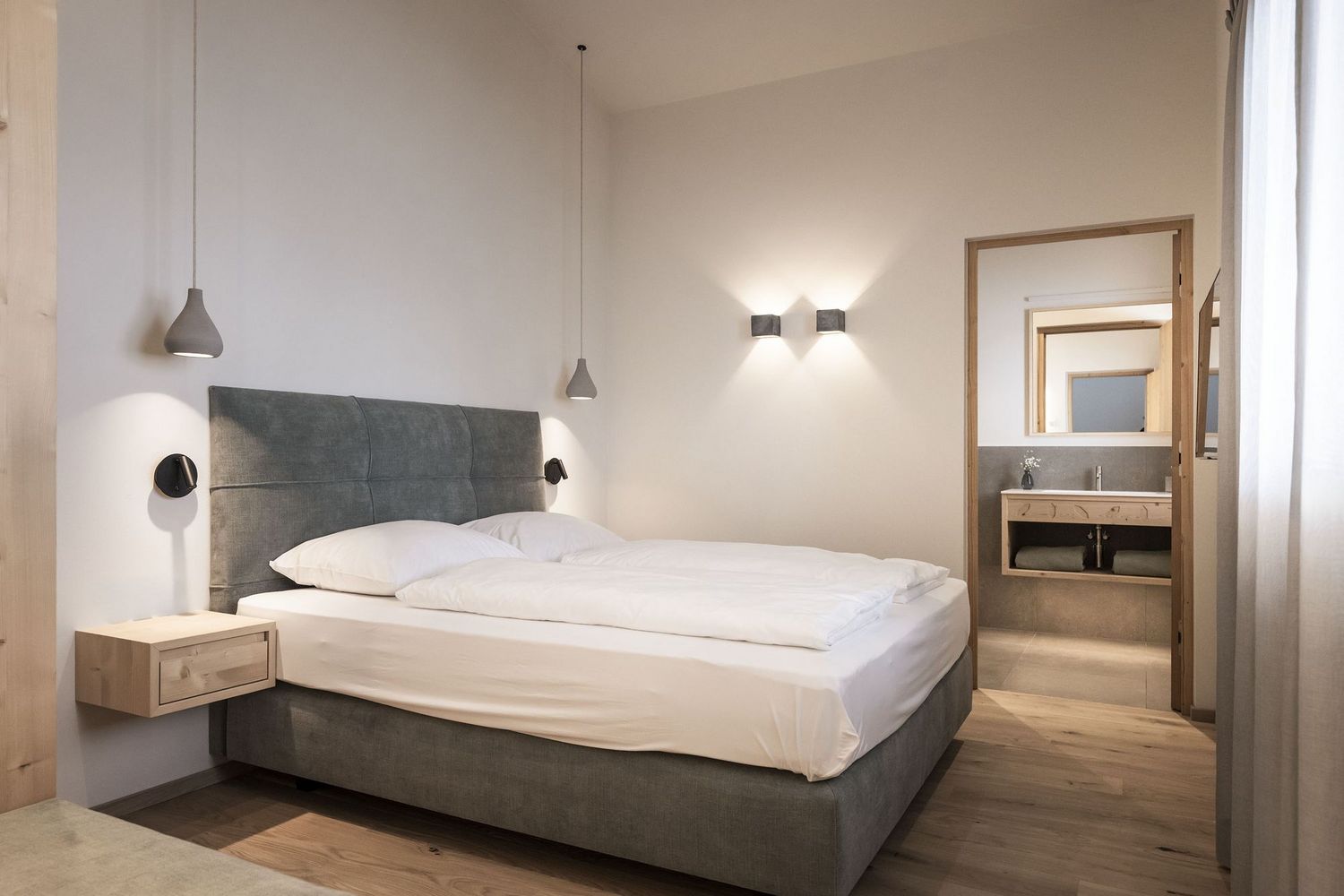 Modernes Schlafzimmer mit grauem Bett, weißen Bettdecken, zwei Hängelampen und Blick in ein angrenzendes Badezimmer
