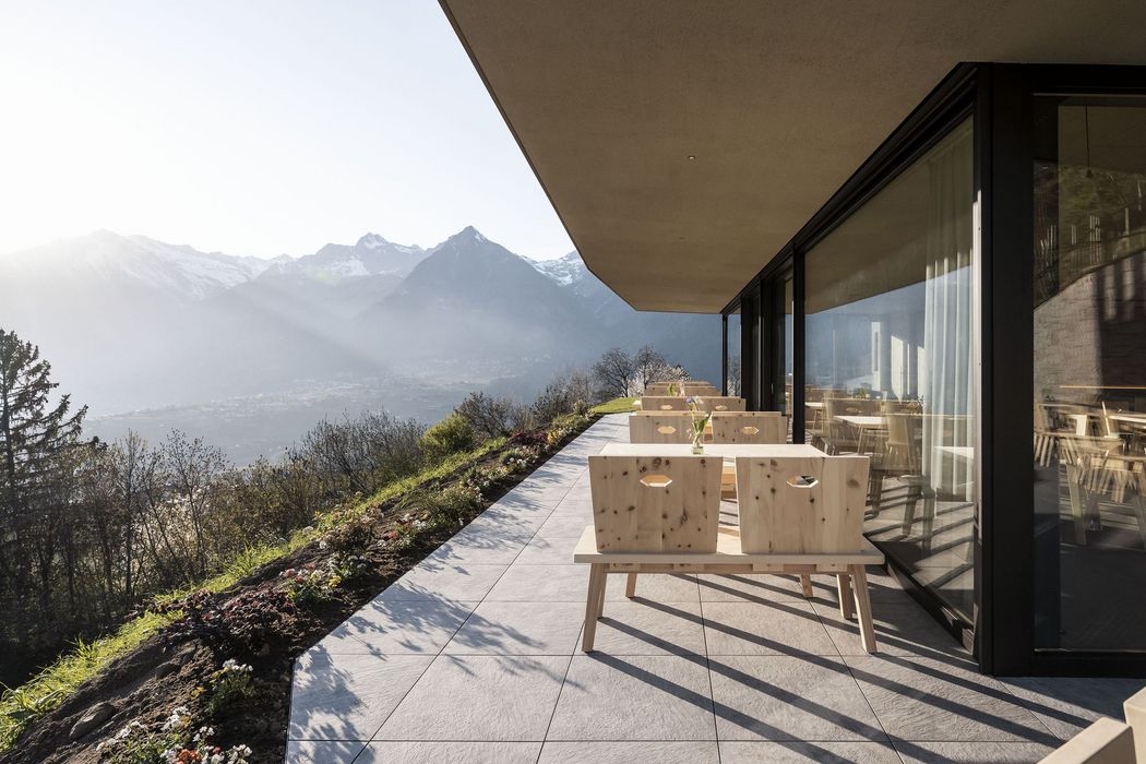 Terrasse mit Holztischen und -bänken neben einem modernen Gebäude mit Blick auf eine Berglandschaft im Sonnenlicht
