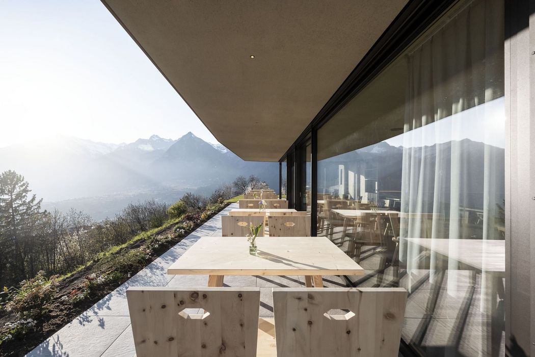 Terrasse mit Holztischen und -stühlen vor Glasfassade, Bergpanorama im Hintergrund bei Sonnenlicht