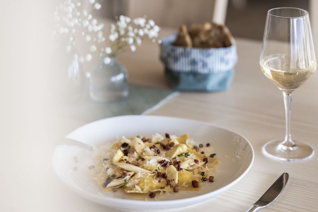 Ein Teller Ravioli mit Speck, Käse und Kräutern auf einem gedeckten Tisch, daneben ein Glas Weißwein.