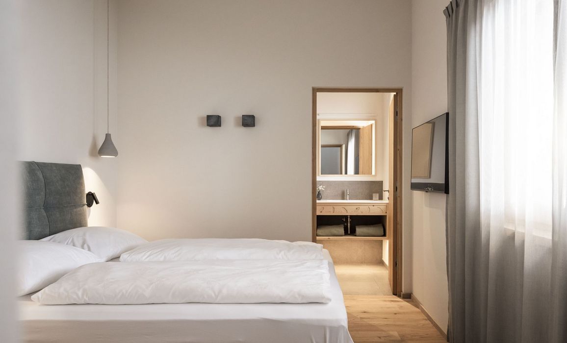 Minimalistisches Schlafzimmer mit weißem Bett, grauem Kopfteil, Hängeleuchte, TV an der Wand und Blick ins Bad