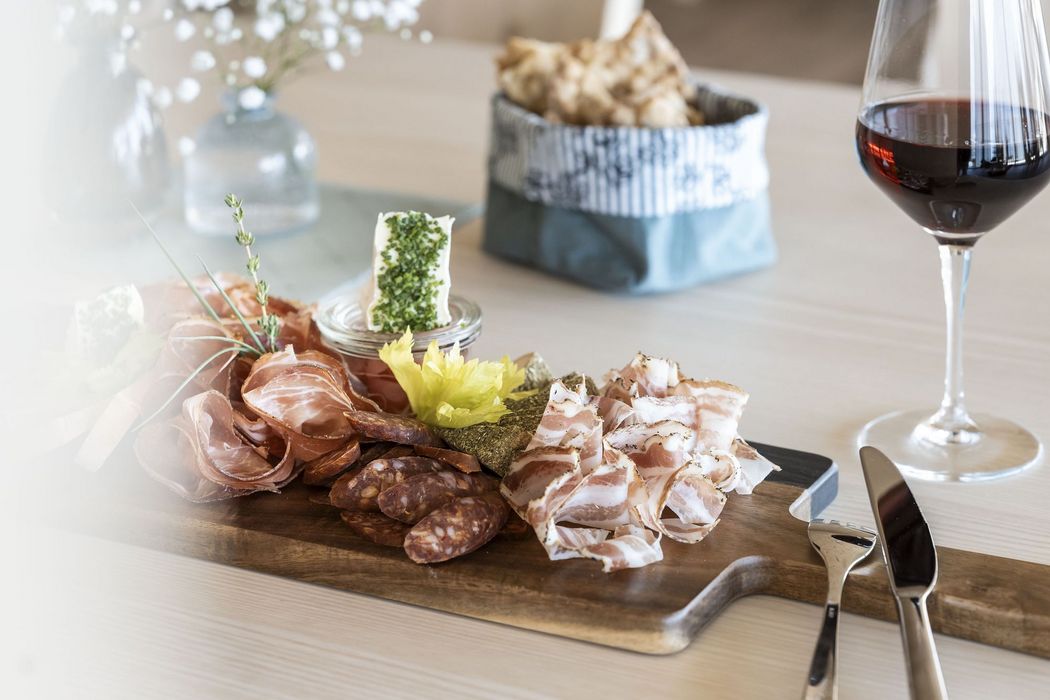 Holzbrett mit verschiedenen Wurst- und Käsesorten, Kräutern, Brot im Hintergrund und Glas Rotwein auf einem Tisch