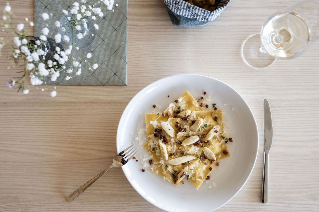 Teller mit Ravioli, Spargelstücken, Käse und Speck auf einem Holztisch mit Besteck, Glas und Blumen in der Vase