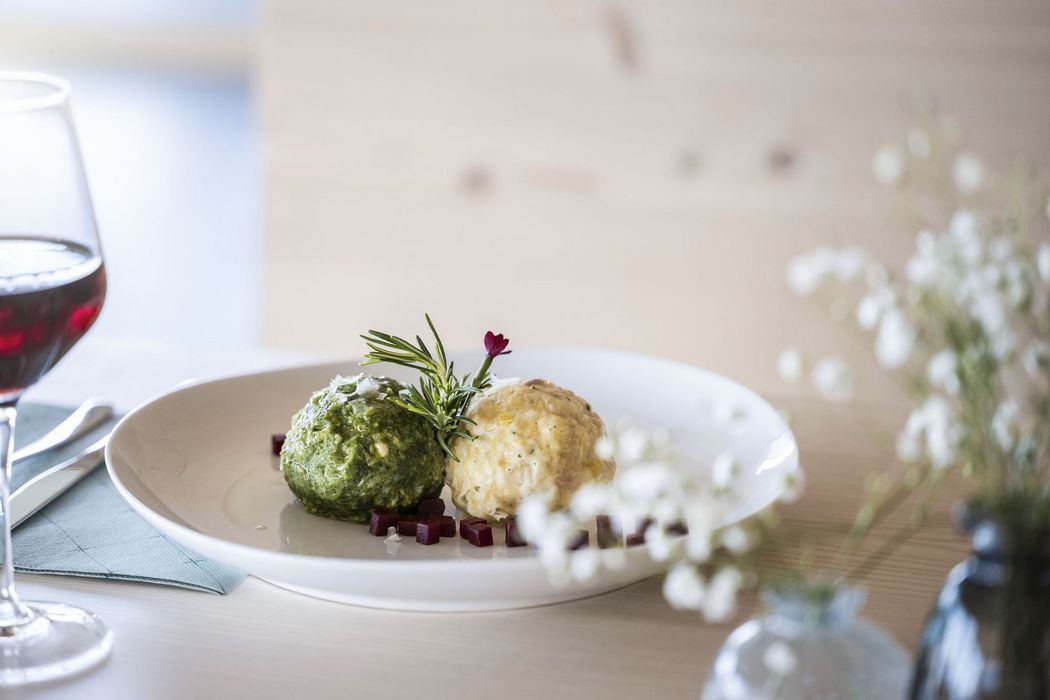 Zwei Kloßbällchen, ein grüner und ein weißer, garniert mit Rosmarin auf einem weißen Teller, daneben ein Glas Rotwein und weiße Blumen.