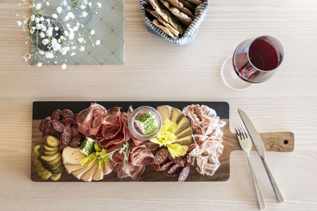 Brett mit verschiedenen Wurst- und Käsesorten, Essiggurken und Kräutercreme, daneben Glas Rotwein und Besteck auf hellem Holztisch