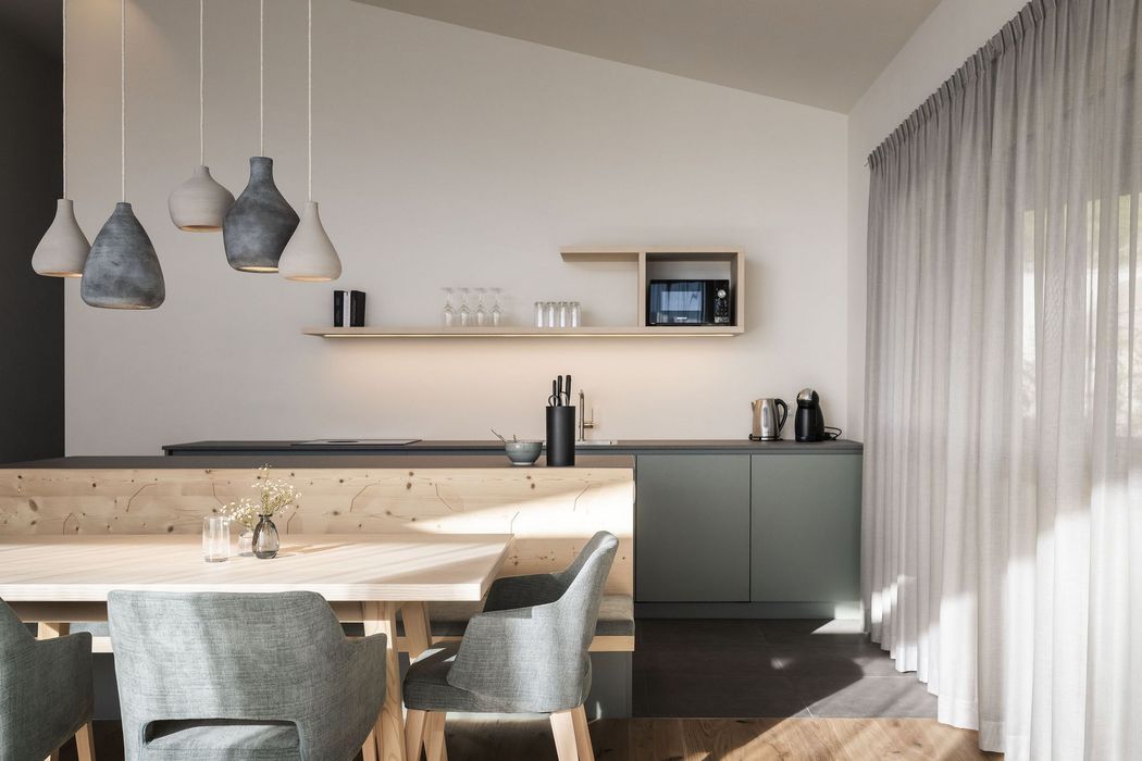 Moderne minimalistische Küche mit Holztisch, grauen Stühlen, Hängelampen und Küchenzeile mit Mikrowelle und Kaffeemaschine.