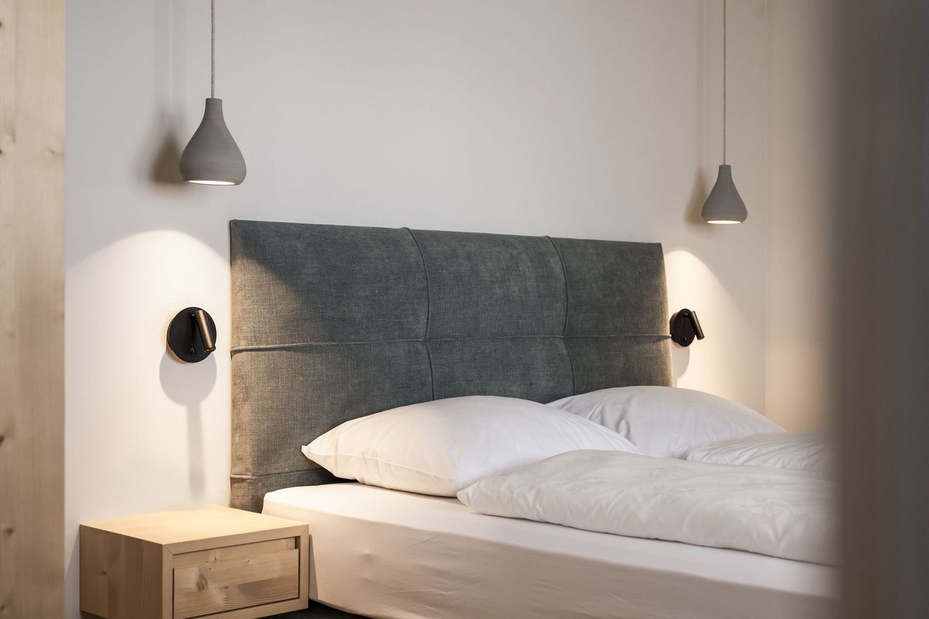 Modernes Schlafzimmer mit grauem gepolstertem Kopfteil, zwei weißen Kissen, Bettdecke, Holz-Nachttisch und zwei hängenden Lampen an der Wand