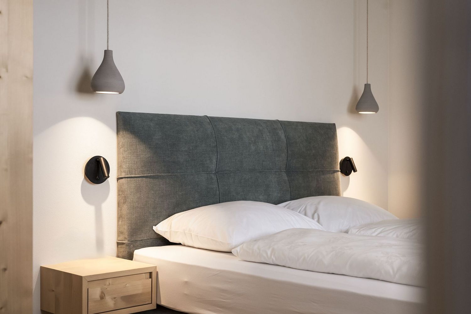 Modernes Schlafzimmer mit grauem gepolstertem Kopfteil, zwei weißen Kissen, Bettdecke, Holz-Nachttisch und zwei hängenden Lampen an der Wand