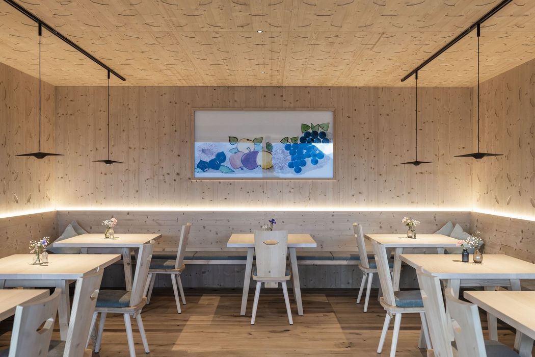 Heller, moderner Gastraum mit Holzwänden, Holztischen und Stühlen, dekorativen Blumenvasen auf den Tischen und einem Bild mit Früchten an der Wand.