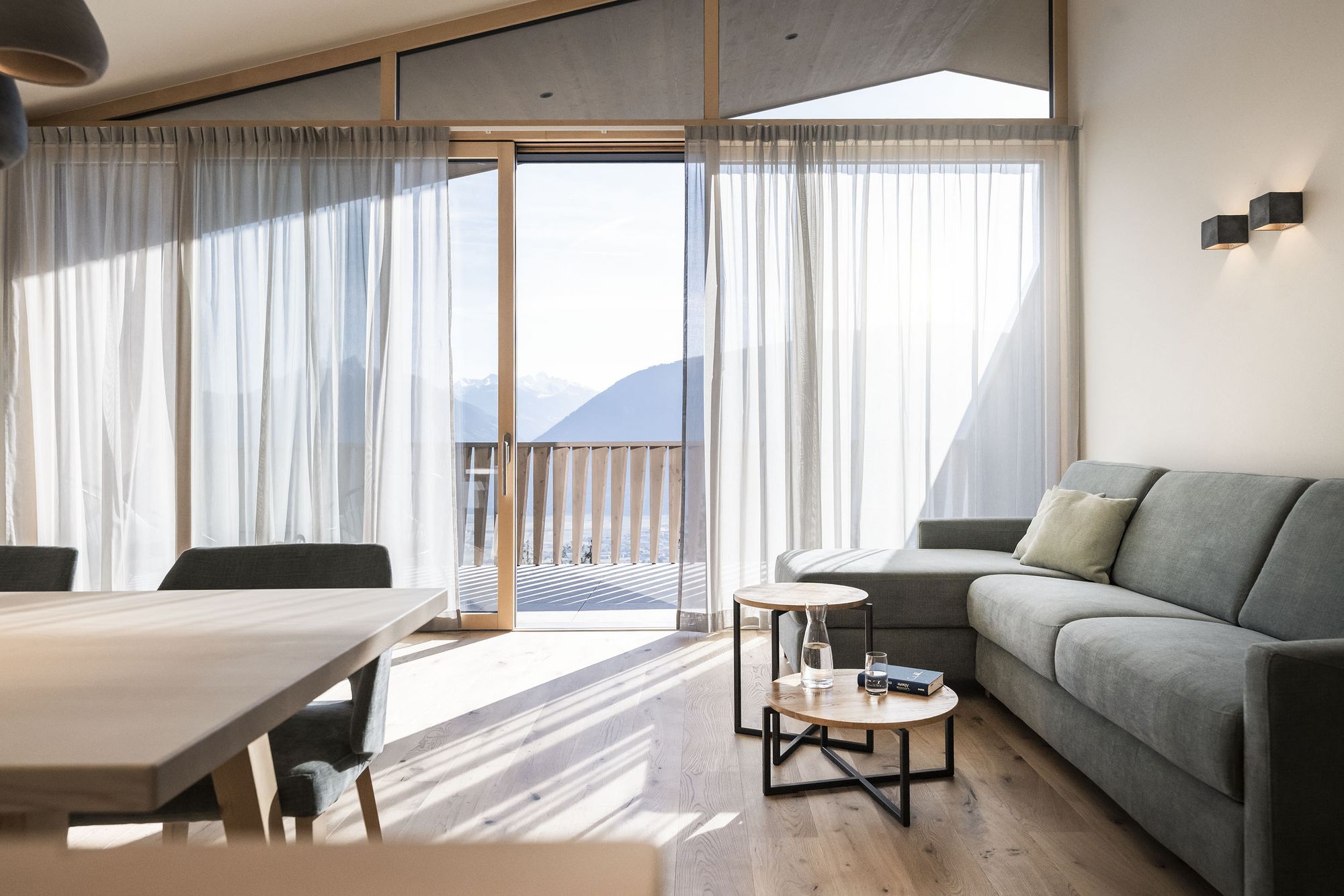Heller, moderner Wohnraum mit großem Sofa, Holztisch, durchscheinenden Vorhängen und Blick auf Berglandschaft durch große Fenster.