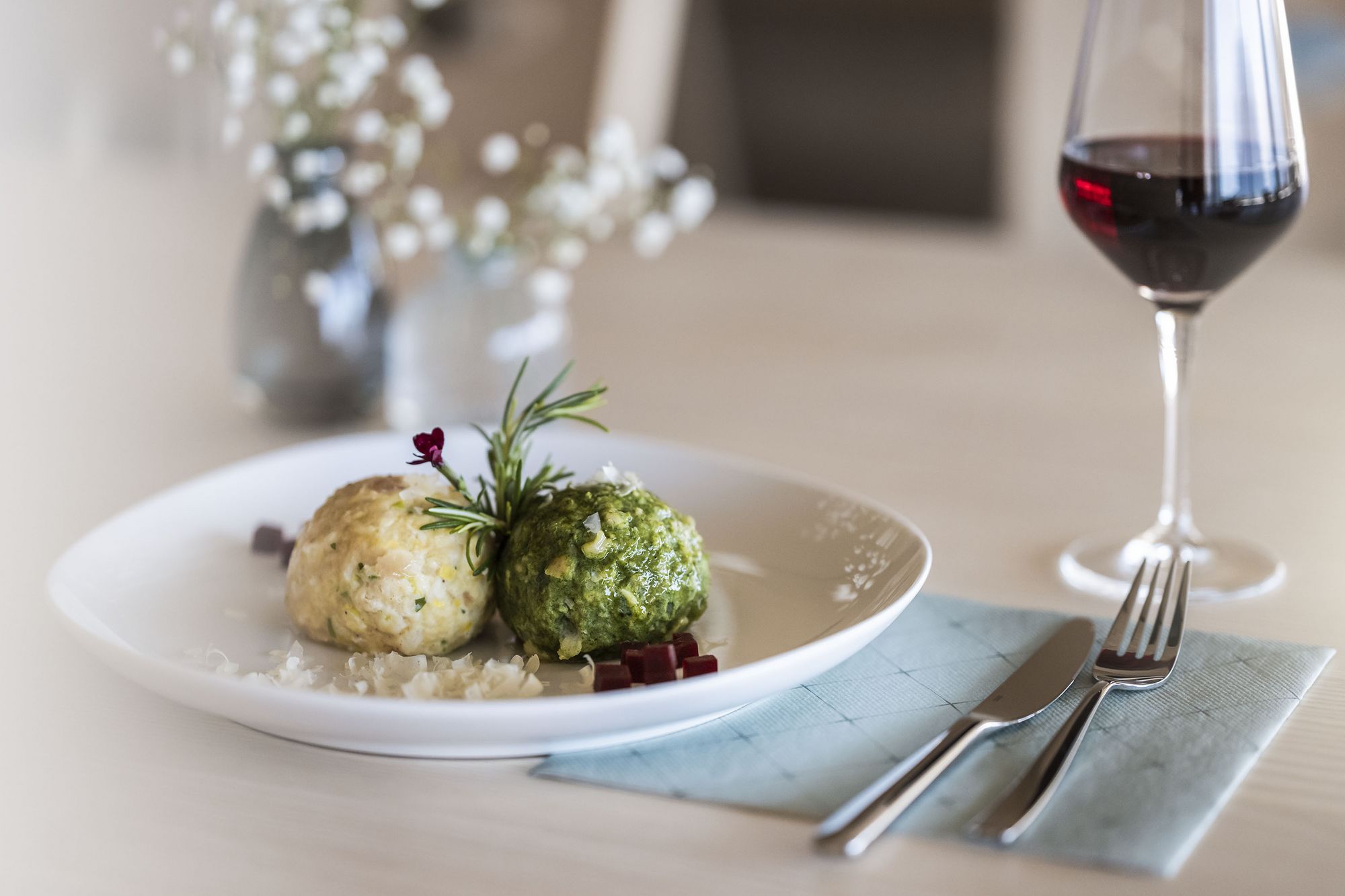 Zwei Knödel auf weißem Teller mit Kräuterzweig, daneben Besteck auf blauer Serviette und ein Glas Rotwein