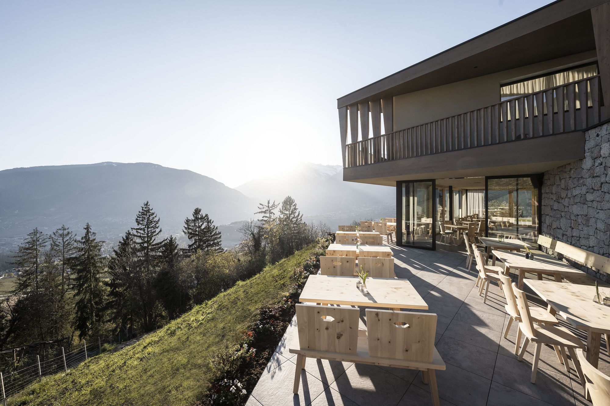 Terrasse mit Holztischen und Stühlen neben modernem Gebäude mit Blick auf Berge und Sonnenuntergang